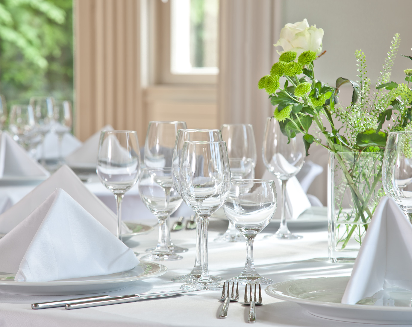 Ein eleganter Tisch ist mit weißen Tischdecken, gefalteten Servietten, Gläsern und Besteck gedeckt. In der Mitte steht eine Vase mit weißen Rosen und grünen Blumen. Im Hintergrund sind Fenster mit Licht erkennbar.