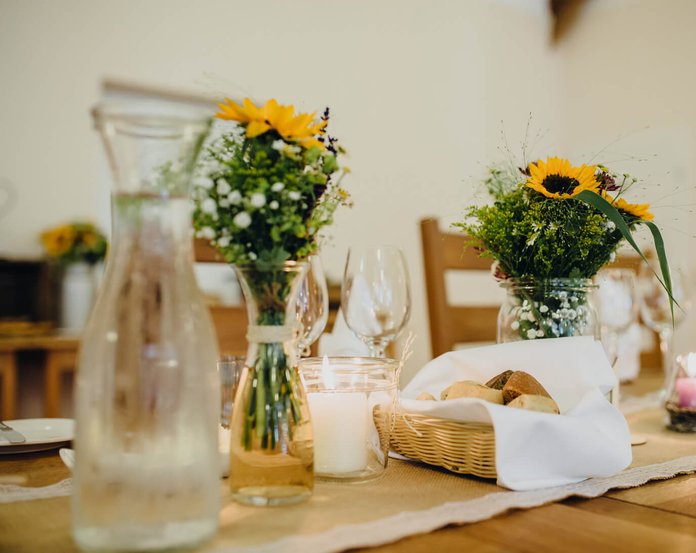 Ein Tisch ist mit Blumensträußen in Gläsern, Kerzen, Wein- und Wassergläsern gedeckt. Ein Korb mit Brot steht auf einem Tischläufer. Die Atmosphäre wirkt warm und einladend.