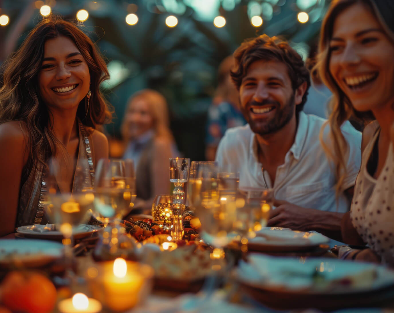 Eine Gruppe von Menschen sitzt an einem Tisch im Freien und lächelt. Vor ihnen stehen beleuchtete Kerzen und Gläser mit Getränken. Im Hintergrund sind Lichterketten und unscharfe Pflanzen zu sehen.